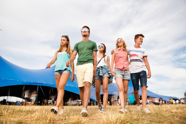 Group of teenage boys and girls at summer music festival walking in front of big blue tent, sunny day