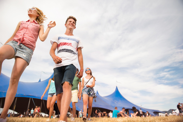 Group of teenage boys and girls at summer music festival walking in front of big blue tent, sunny day