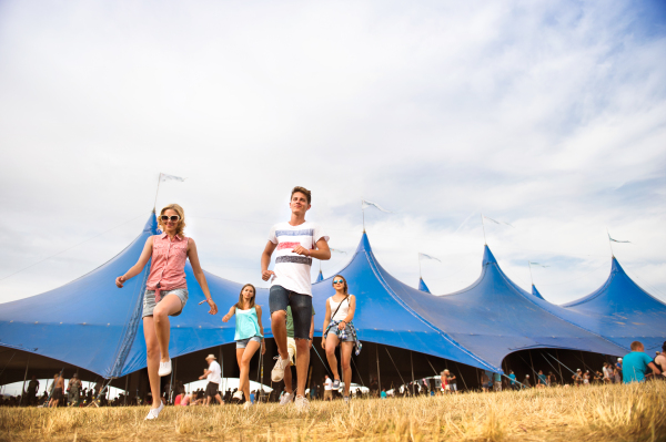 Group of teenage boys and girls at summer music festival walking in front of big blue tent, sunny day