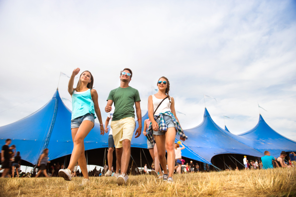 Group of teenage boys and girls at summer music festival walking in front of big blue tent, sunny day