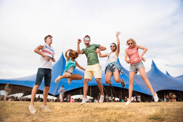 Group of teenage boys and girls at summer music festival, dancing and jumping in front of big tent, sunny day