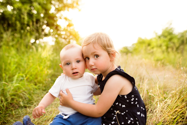 Cute little blonde girl in blue dress holding her baby brother outside in in green sunny summer nature