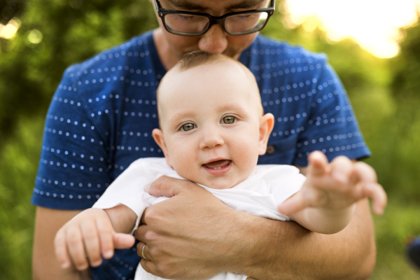 Young father in green summer nature holding his cute baby son in the arms, kissing him.