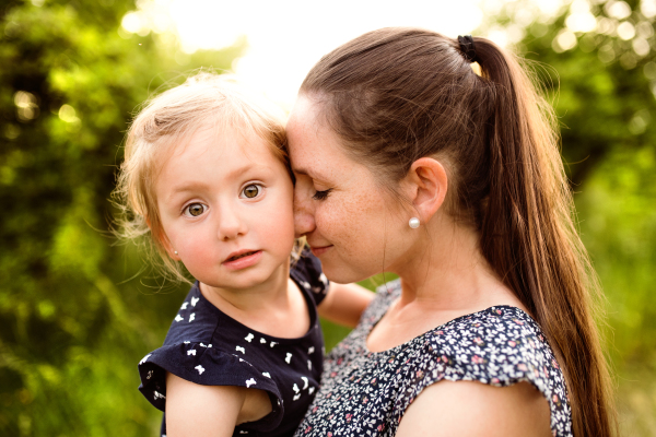 Beautiful young mother in green sunny summer nature holding her cute little daughter in the arms.