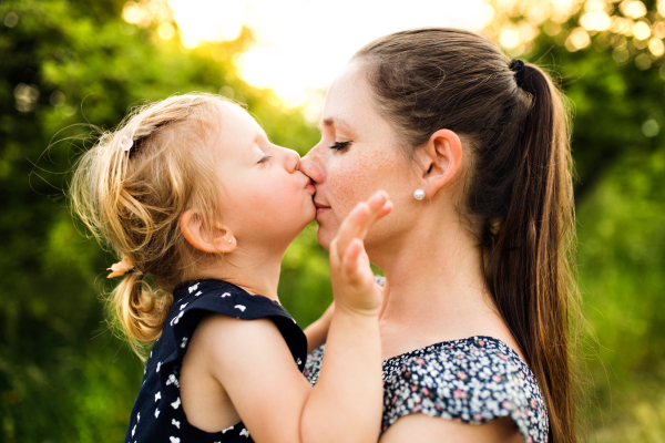 Beautiful young mother in green sunny summer nature holding her cute little daughter in the arms, girl kissing her on nose.