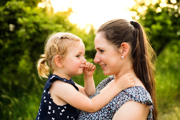 Beautiful young mother in green sunny summer nature holding her cute little daughter in the arms.