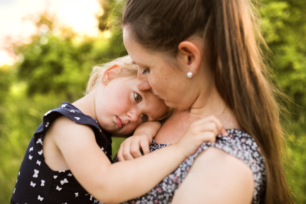 Beautiful young mother in green sunny summer nature holding her cute little daughter in the arms.