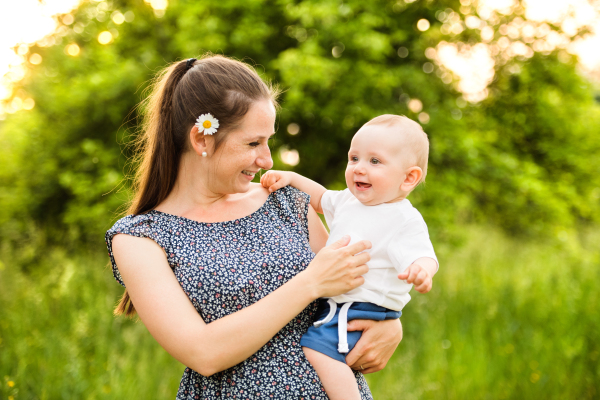 Beautiful young mother in green sunny summer nature holding baby son in the arms.