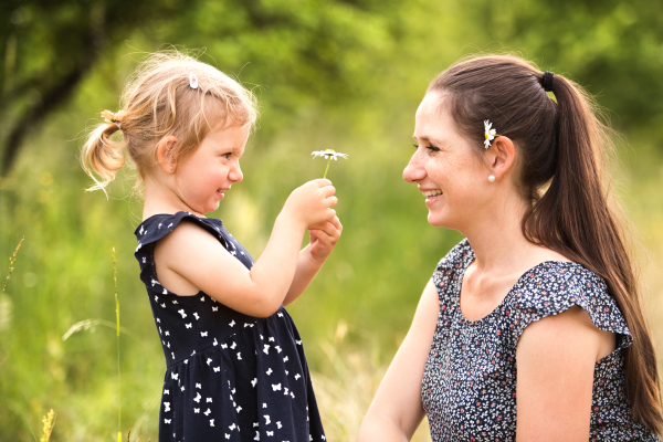 Beautiful young mother in green sunny summer nature with her cute little daughter, girl giving her daisy flower.