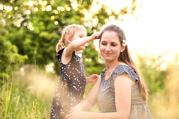 Beautiful young mother in green sunny summer nature with her cute little daughter, girl giving her daisy flower.