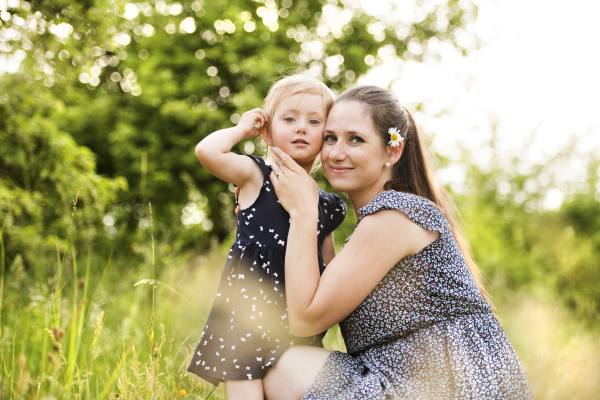 Beautiful young mother in green sunny summer nature holding her cute little daughter in the arms.