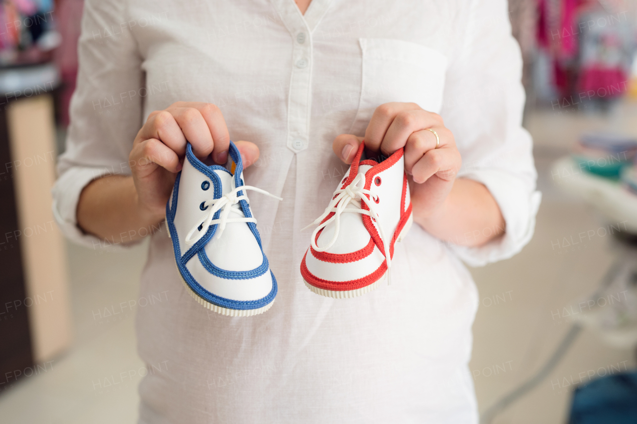 Unrecognizable pregnant woman in white shirt shopping shoes for her baby, not knowing whether to buy blue for boy or red for girl