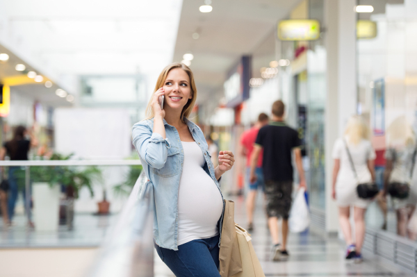 Beautiful young pregnant woman holding smart phone, making phone call, in shopping center, resting