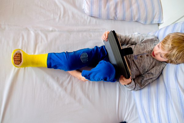 Little boy with broken leg lying in bed playing on tablet