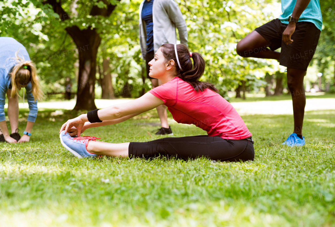 Group of fit runners in nature warming up and stretching. Beautiful young woman sitting on the grass stretching her legs and arms.