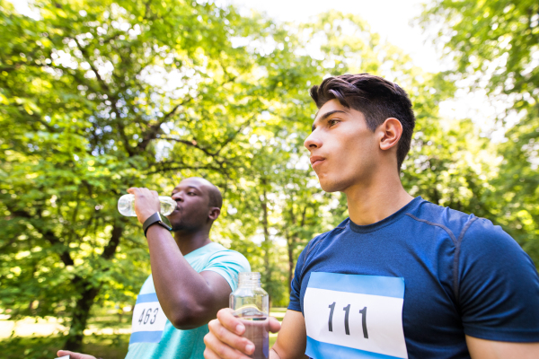 Two young athletes prepared for run in green sunny summer park, holding bottles, drinking water.