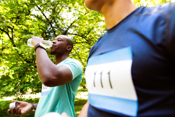 Two young athletes prepared for run in green sunny summer park, holding bottles, drinking water.