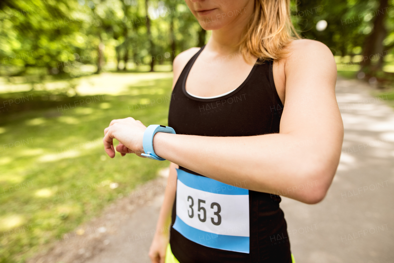 Unrecognizable young woman setting up watch before going for a run in green sunny summer park.