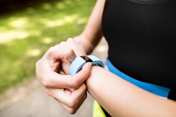 Unrecognizable young woman setting up watch before going for a run in green sunny summer park.