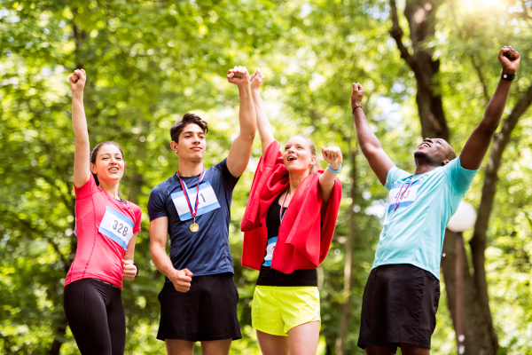 Group of young fit friends at the finish line happy after completing race, arms raised. Green sunny park.