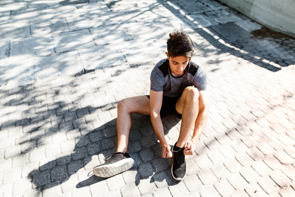 Young hispanic runner in the city sitting on concrete path tying his shoelaces.