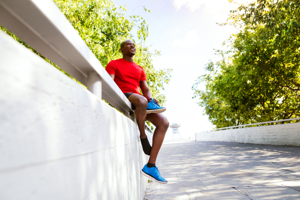 Young afro-american runner in the city sitting on concrete wall holding a shoe, resting.