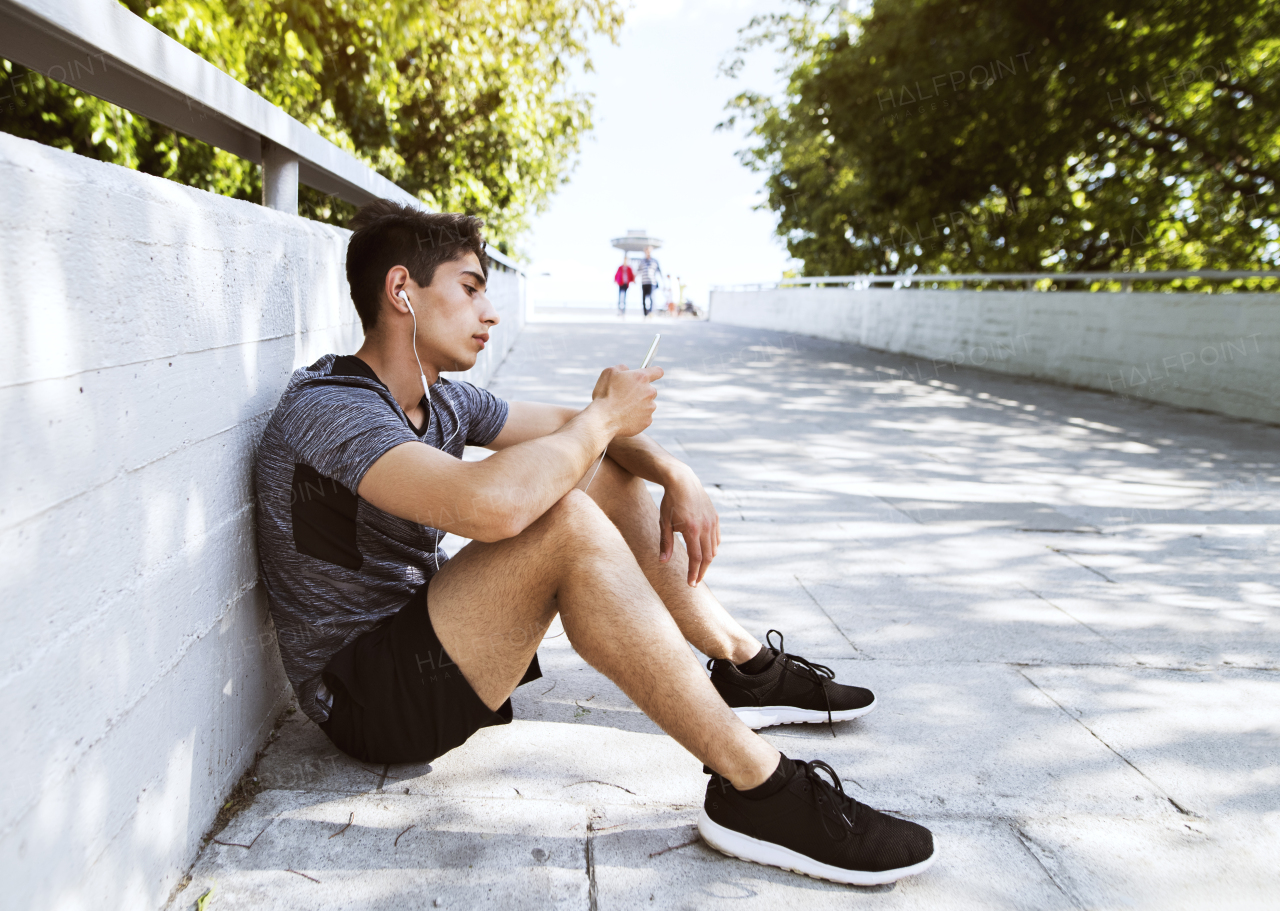 Handsome young runner with smartphone in the city park, resting, earphones in his ears, listening to music.