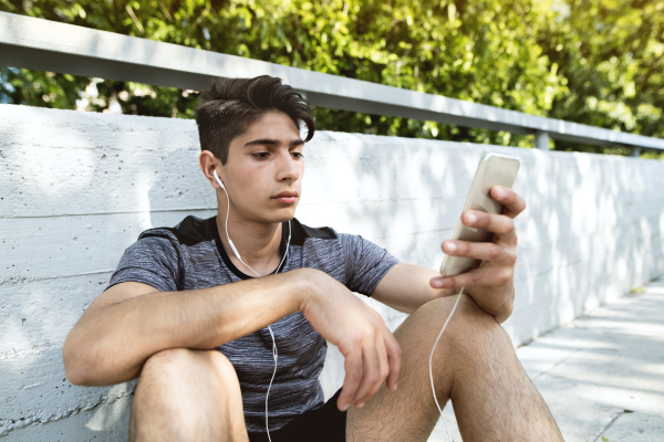 Handsome young runner with smartphone in the city park, resting, earphones in his ears, listening to music.