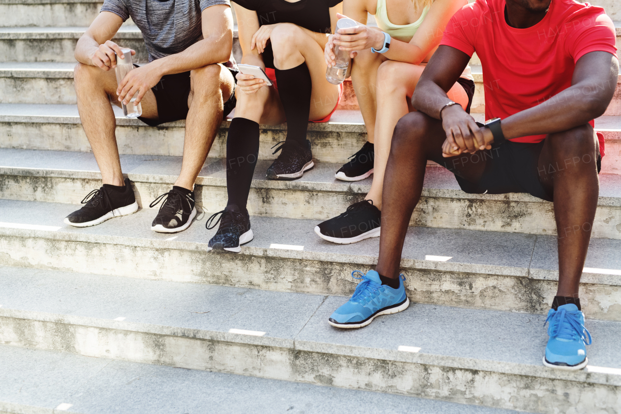 Unrecognizable young runners in the city together sitting on the stairs, looking at smart watch or smart phone, tracking their progress in running.