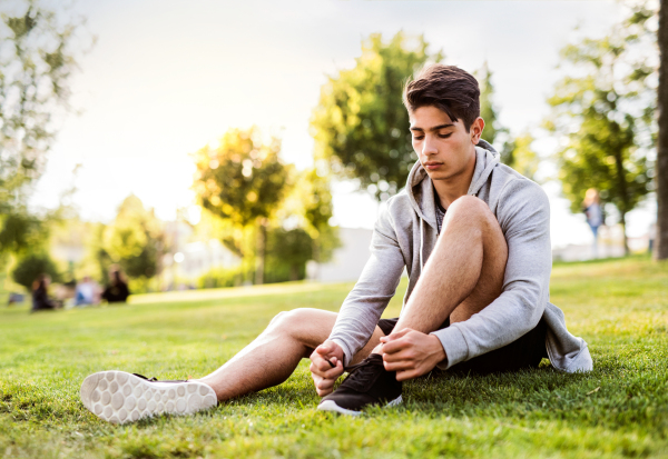 Young hispanic runner in the city sitting on the grass in park tying his shoelaces.