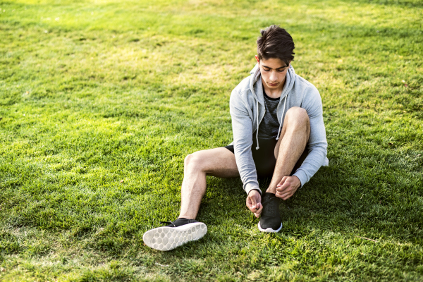 Young hispanic runner in the city sitting on grass tying his shoelaces.