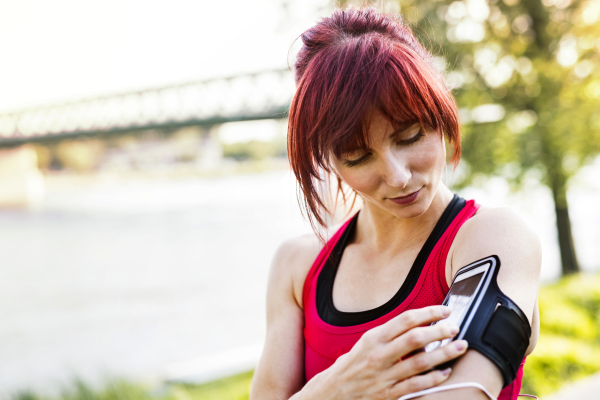 Beautiful young runner in sports clothes running in the city park, resting. Woman with smartphone in an armband.