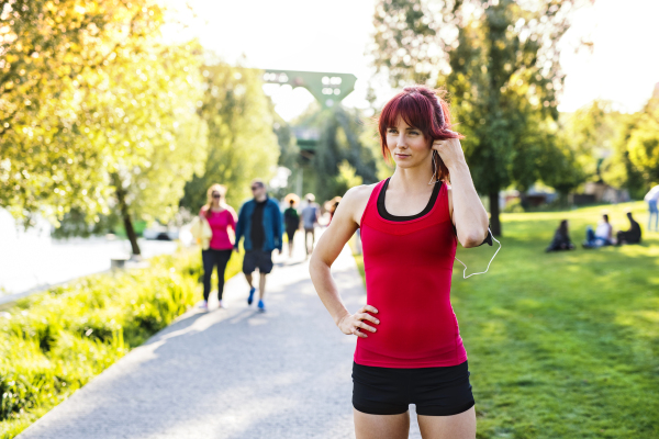 Beautiful young runner in sports clothes running in the city park, resting. Woman with earphones listening to music.