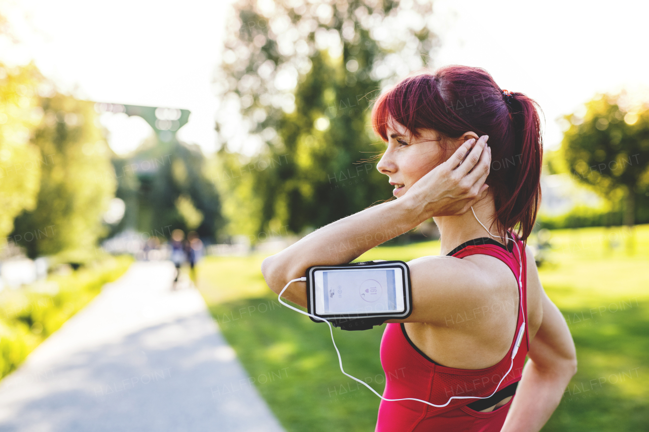 Beautiful young runner in sports clothes running in the city park, resting. Woman with earphones listening to music. Smartphone in an armband.