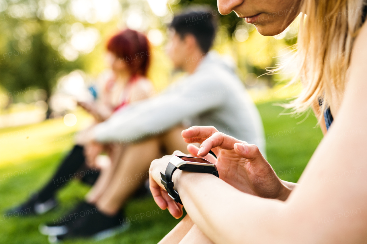 Unrecognizable young runner in park, sitting on grass, setting her smartwatch.