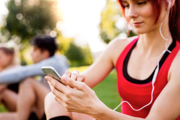 Beautiful young runner in park, sitting on grass, holding smart phone, listening music.