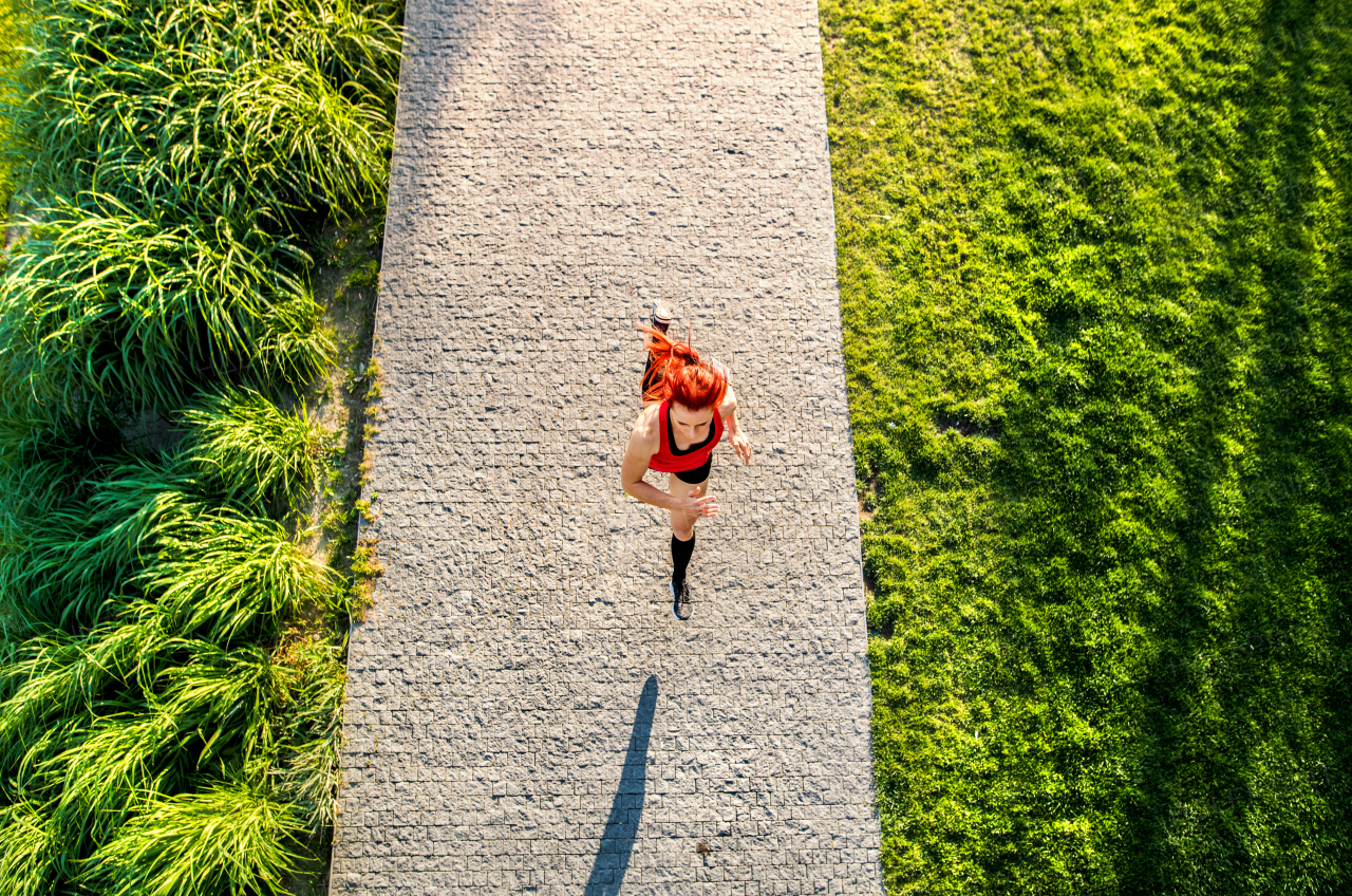 Beautiful young athlete in sports clothes running on concrete path in green sunny park.