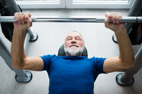 Senior man in sports clothing in gym working out with weights, bench pressing.