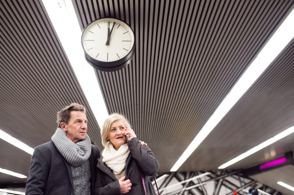 Beautiful senior couple standing at the underground platform, waiting. Woman with smart phone making phone call.