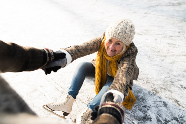 Beautiful senior couple in sunny winter nature ice skating, unrecognizable man helping woman to stand up.