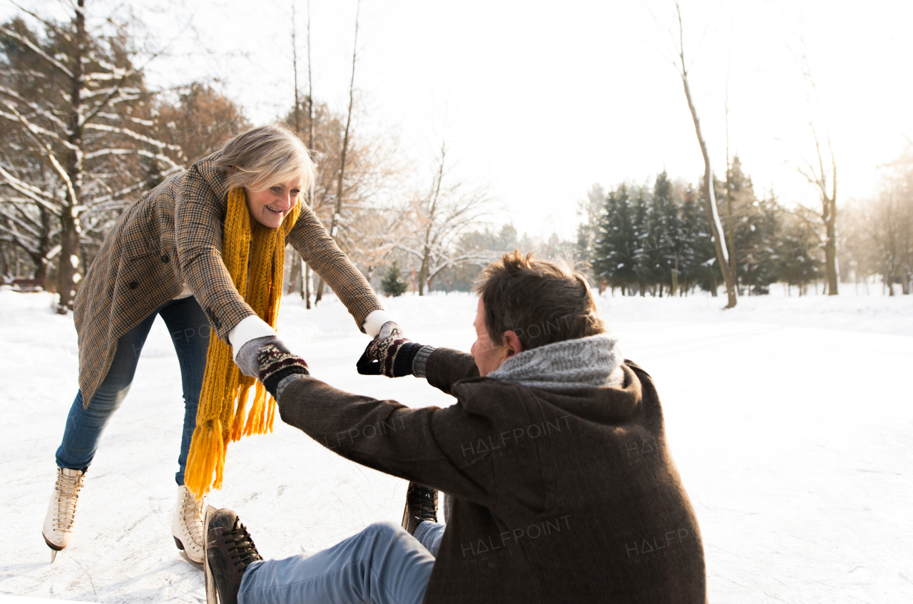 Beautiful senior couple in sunny winter nature ice skating, woman helping man to stand up.