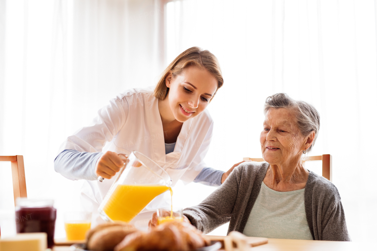 Health visitor and a senior woman during home visit. A nurse pouring orange juice to and an elderly woman.