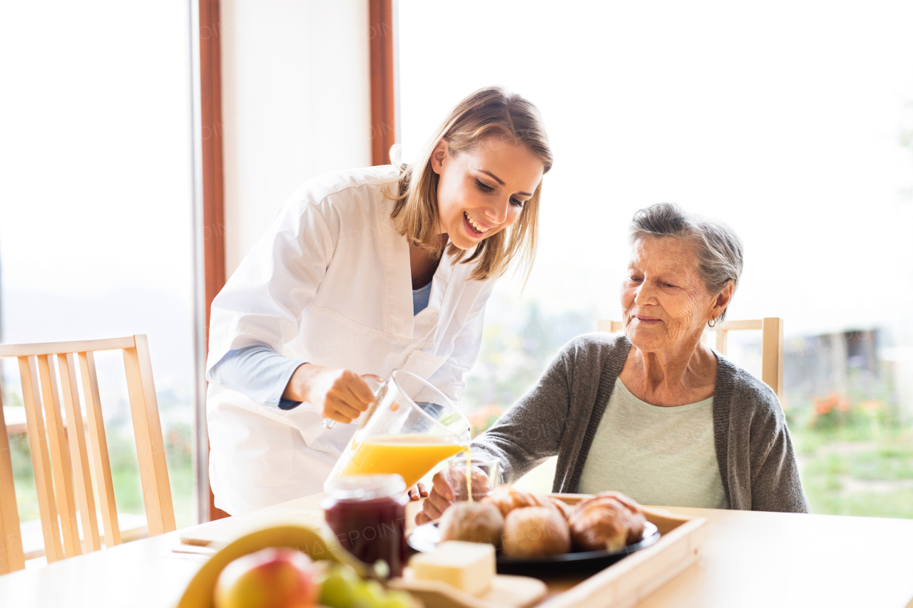 Health visitor and a senior woman during home visit. A nurse pouring orange juice to an elderly woman.