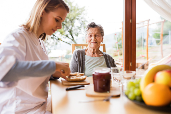 Health visitor and a senior woman during home visit. A nurse and an elderly woman sitting at the table, preparing breakfast.