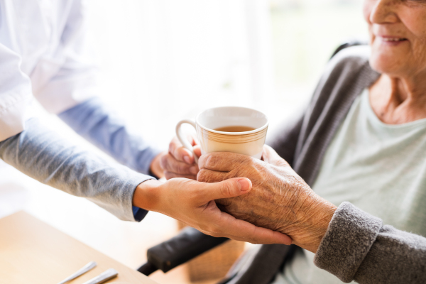 Unrecognizable health visitor and a senior woman during home visit. A nurse giving a cup of tea to an elderly woman sitting at the table. Close up.