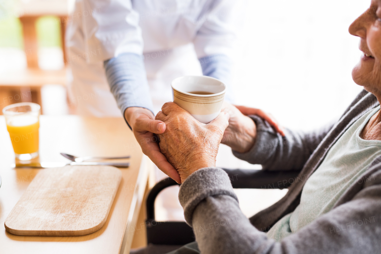 Unrecognizable health visitor and a senior woman during home visit. A nurse giving tea to an elderly woman sitting at the table. Close up.