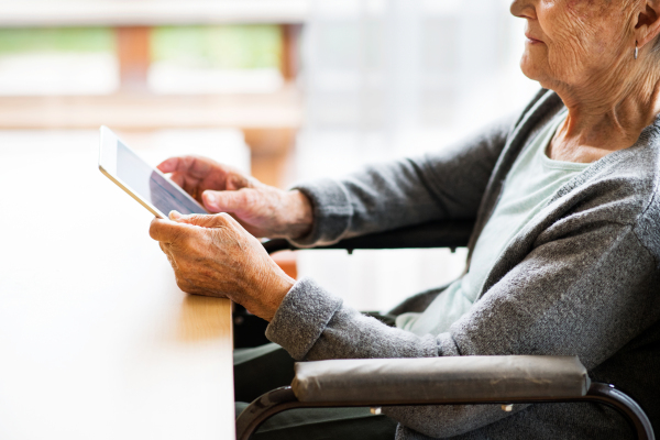 Unrecognizable senior woman in a wheelchair with tablet at home. An elderly woman sitting at the table.