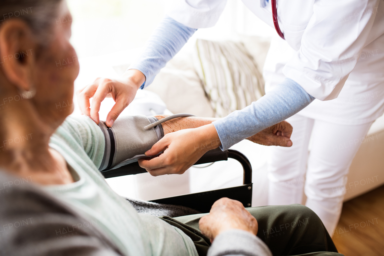 Unrecognizable health visitor and a senior woman during home visit. A nurse checking blood pressure of a woman in an wheelchair.