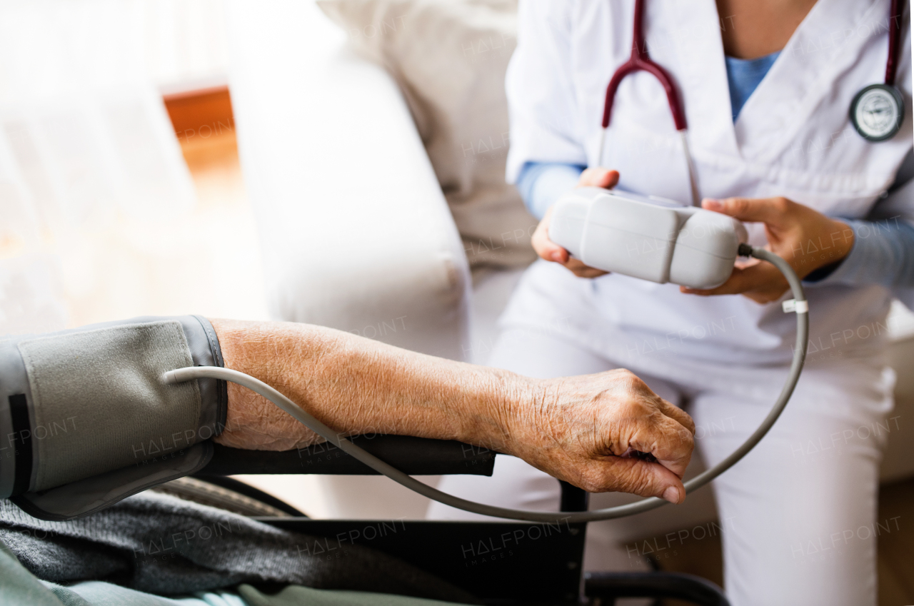 Unrecognizable health visitor and a senior woman during home visit. A nurse checking blood pressure of a woman in an wheelchair.