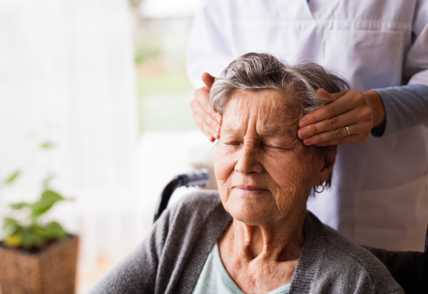 Health visitor and a senior woman during home visit. Unrecognizable nurse giving woman temple massage.